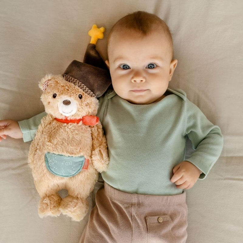 Baby lying on a beige surface with a teddy bear wearing a hat